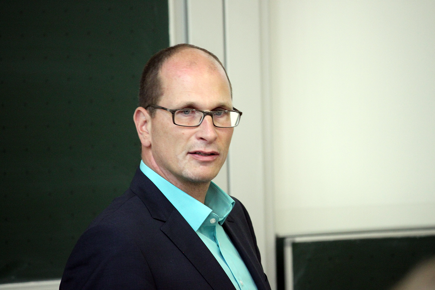 Ein Mann in dunklem Anzug und türkisem Hemd, der eine Brille trägt, spricht selbstbewusst. Er steht vor einer Tafel und einem Whiteboard und verkörpert den akademischen Geist der Hochschule Coburg im Unterrichtsumfeld.