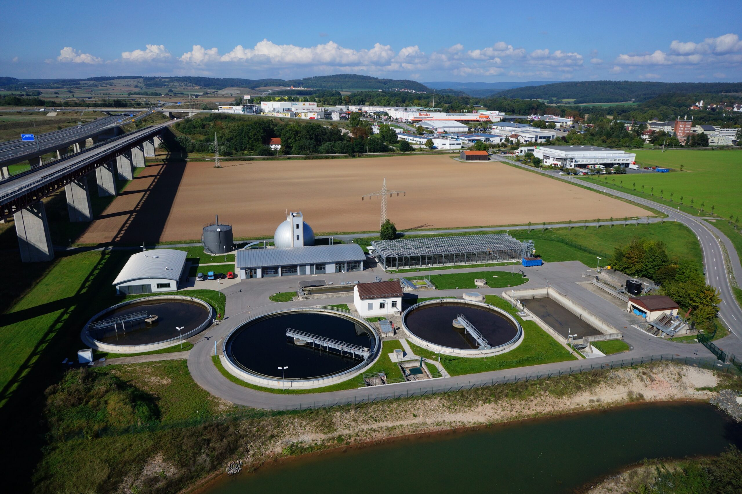 Luftaufnahme der Kläranlage in der Nähe der Hochschule Coburg mit runden Tanks und angrenzenden Gebäuden. Eine Autobahnüberführung verläuft daneben, mit offenen Feldern und entfernten Hügeln im Hintergrund unter einem teilweise bewölkten Himmel.