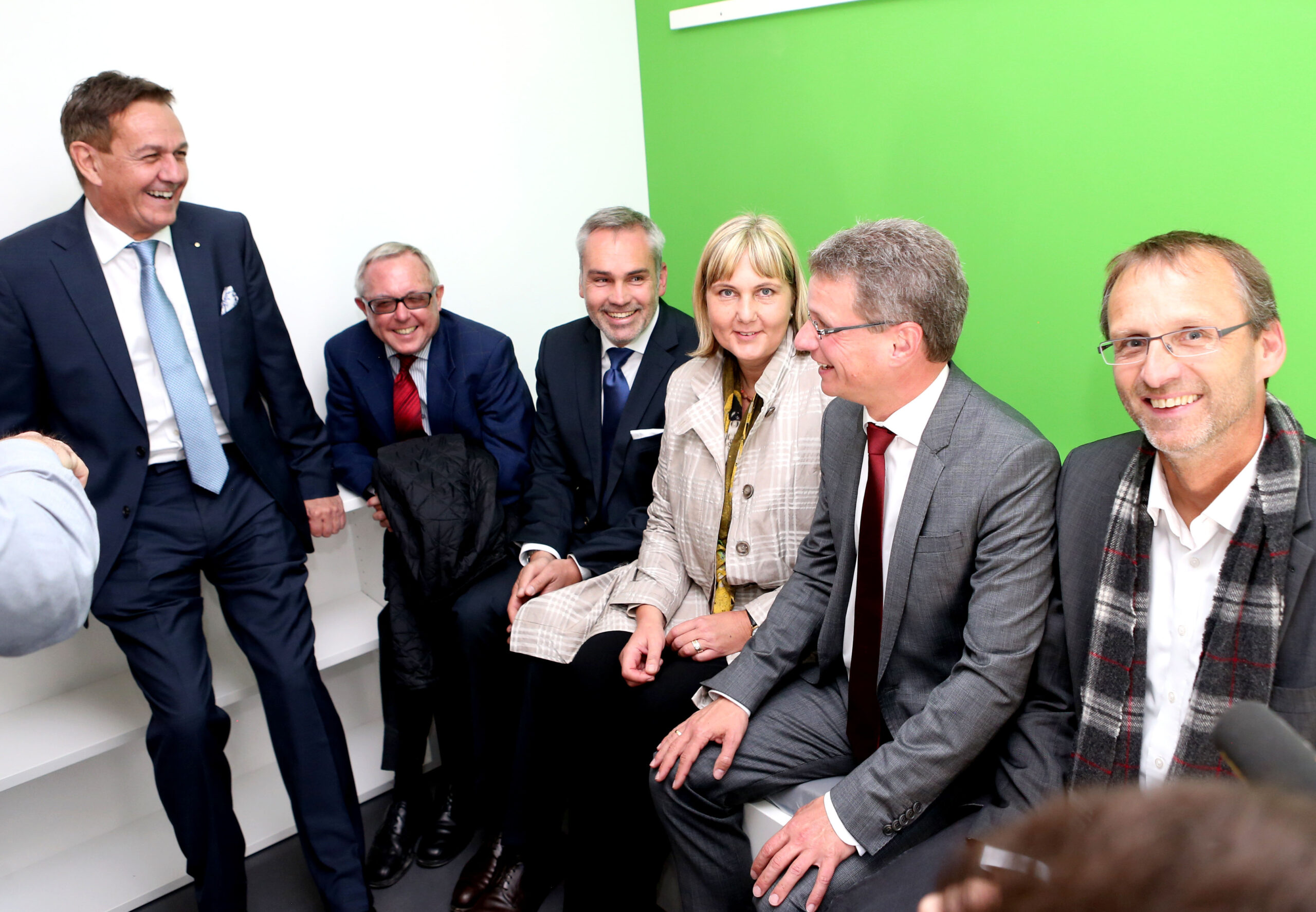 Eine Gruppe von sechs Personen, fünf Männer und eine Frau, sitzt lächelnd zusammen in einem Raum mit grün-weißer Wand der Hochschule Coburg. Sie tragen Business-Kleidung, ein Mann trägt ein Kopftuch.