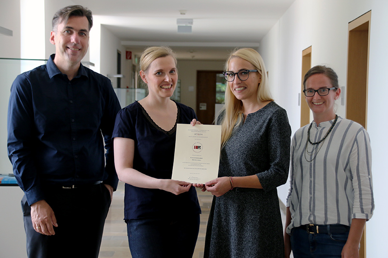 Vier Personen stehen in einem Flur der Hochschule Coburg. Zwei Frauen in der Mitte halten lächelnd ein Zertifikat in den Händen. Die anderen beiden (ein Mann und eine Frau) stehen daneben und lächeln ebenfalls. Die Szenerie scheint ein Büro oder eine Bildungseinrichtung zu sein.