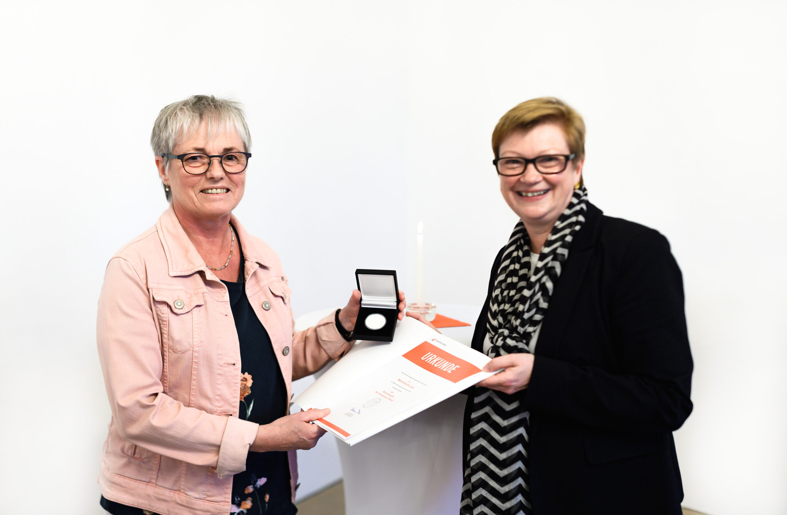 Zwei Personen stehen nebeneinander und lächeln. Eine hält eine Urkunde und eine Medaille in einer offenen Schachtel. Der schlichte weiße Hintergrund lässt vermuten, dass sie an einer Preisverleihung oder einer ähnlichen Veranstaltung teilnehmen, bei der möglicherweise Leistungen an der Hochschule Coburg gewürdigt werden.