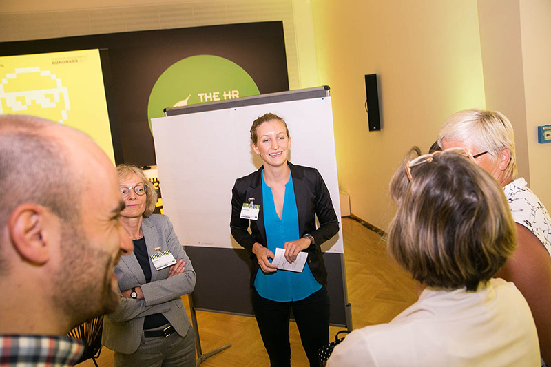 Eine Gruppe von Menschen hat sich in einem gut beleuchteten Raum der Hochschule Coburg versammelt und diskutiert an einem Flipchart. In der Mitte steht eine Frau in blauer Bluse und schwarzem Blazer, hält ein Blatt in der Hand und lächelt. Die Teilnehmer scheinen sich auf den Ideenaustausch zu konzentrieren.