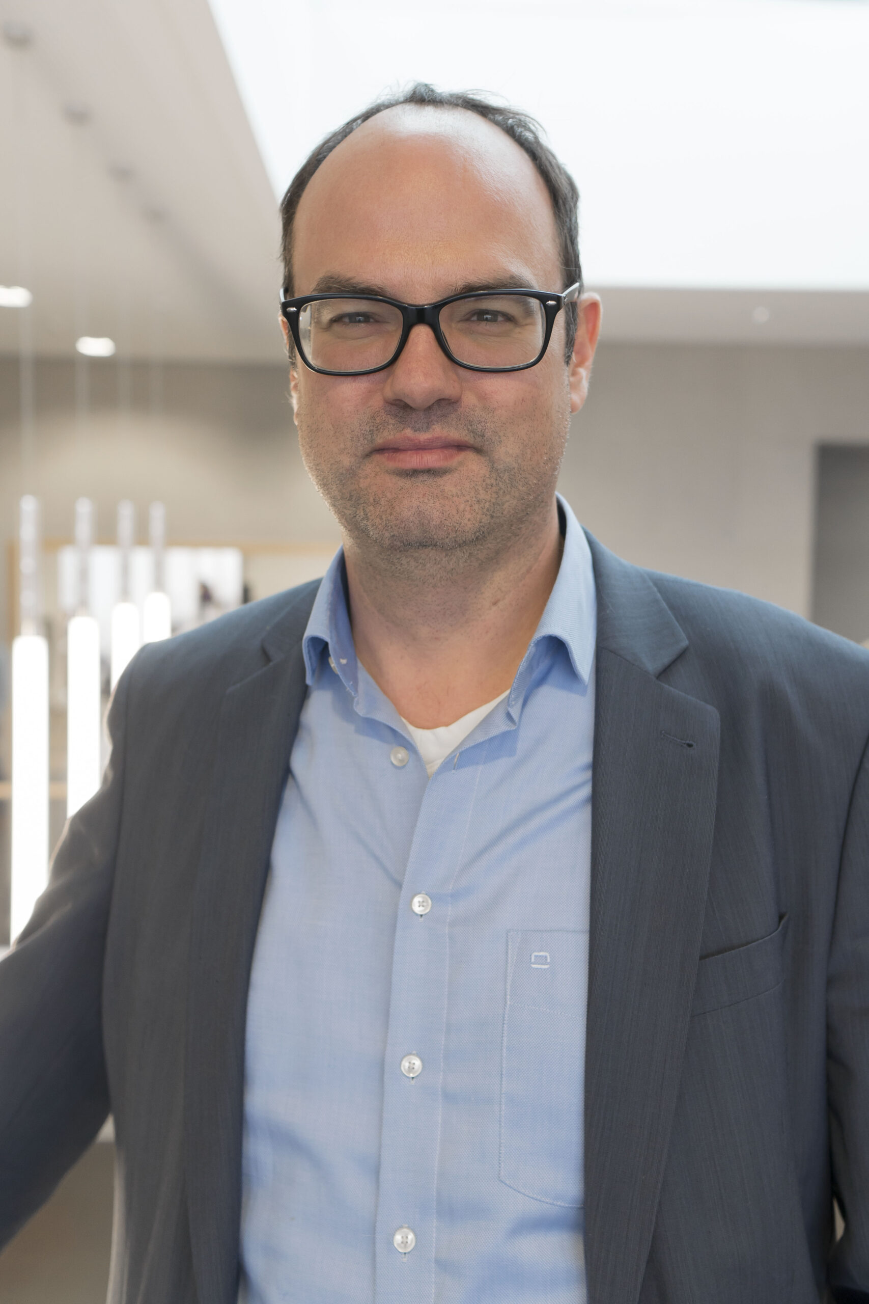 Ein Mann mit Brille, einem blauen Hemd und einem grauen Blazer steht in einem Innenbereich vor einem unscharfen Hintergrund, der an die modernen Campusgelände der Hochschule Coburg erinnert.