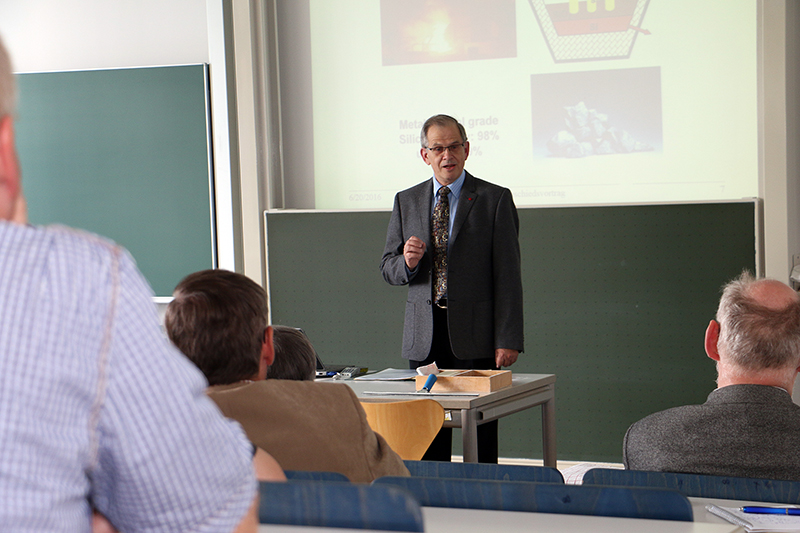 Eine Person hält in einem Hörsaal der Hochschule Coburg eine Präsentation. Sie steht vor einer Tafel und projiziert Folien und spricht zu den sitzenden Teilnehmern. Der Raum ist mit den typischen Möbeln eines Hörsaals ausgestattet und die Kleidung ist formell, was dem akademischen Umfeld entspricht.