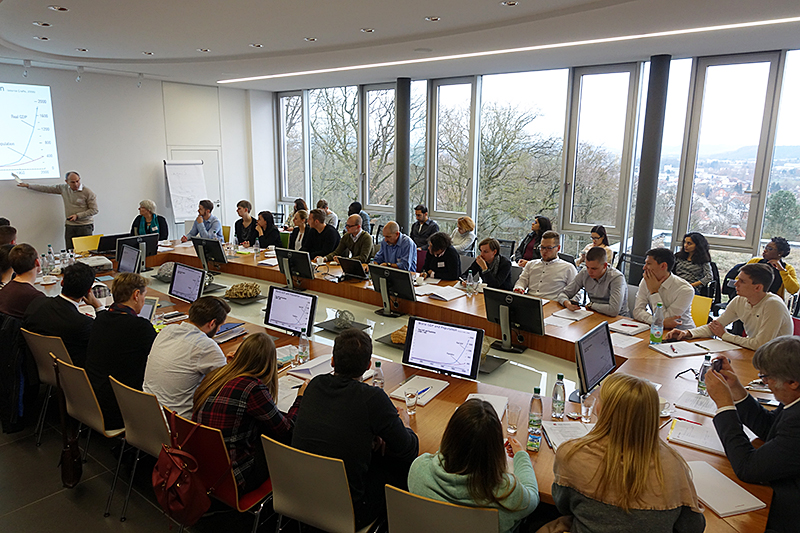 In einem Konferenzraum der Hochschule Coburg sitzt eine Gruppe von Menschen mit Papieren und Monitoren an rechteckigen Tischen. Große Fenster geben den Blick auf die Bäume draußen frei. Vorne steht ein Redner neben einem Whiteboard und spricht zu den Teilnehmern.