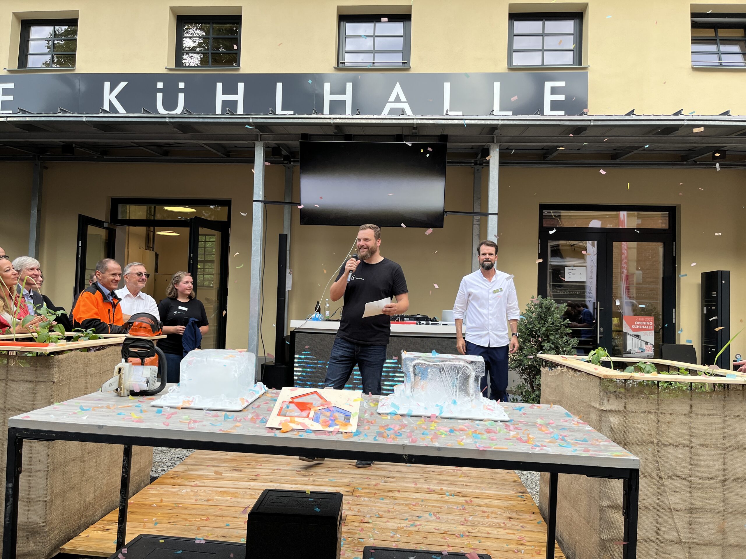 Ein Mann von der Hochschule Coburg spricht in ein Mikrofon auf einer Holzplattform. Auf dem Tisch davor stehen eine Eisskulptur und ein schmelzender Eisblock. Die Leute stehen herum und schauen zu, wie Konfetti von oben herabregnet. Am Veranstaltungsort hängt ein Schild mit der Aufschrift „KÜHLHALLE“.