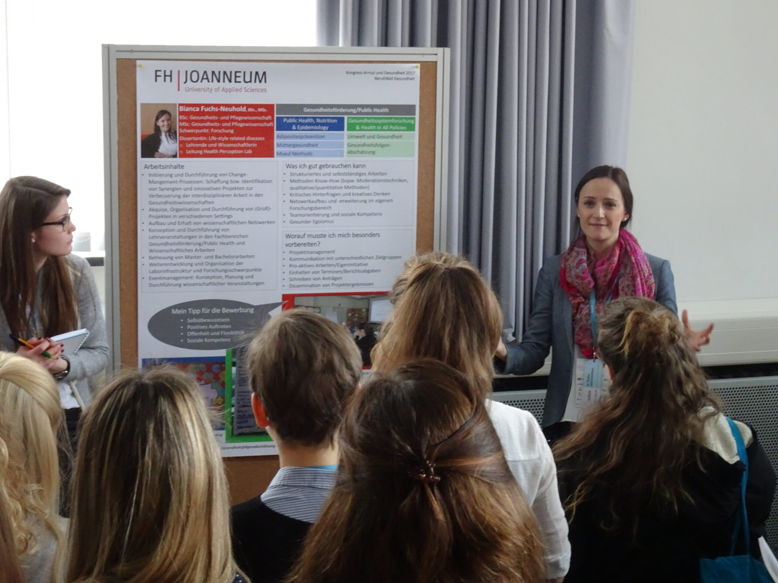 Eine Frau steht neben einem Präsentationsplakat mit dem Titel „FH JOANNEUM“ und unterhält sich mit einer Gruppe. Eine andere Frau, möglicherweise zu Besuch von der Hochschule Coburg, hält einen Notizblock neben sich. Das Publikum hört in der Unterrichtssituation aufmerksam zu.