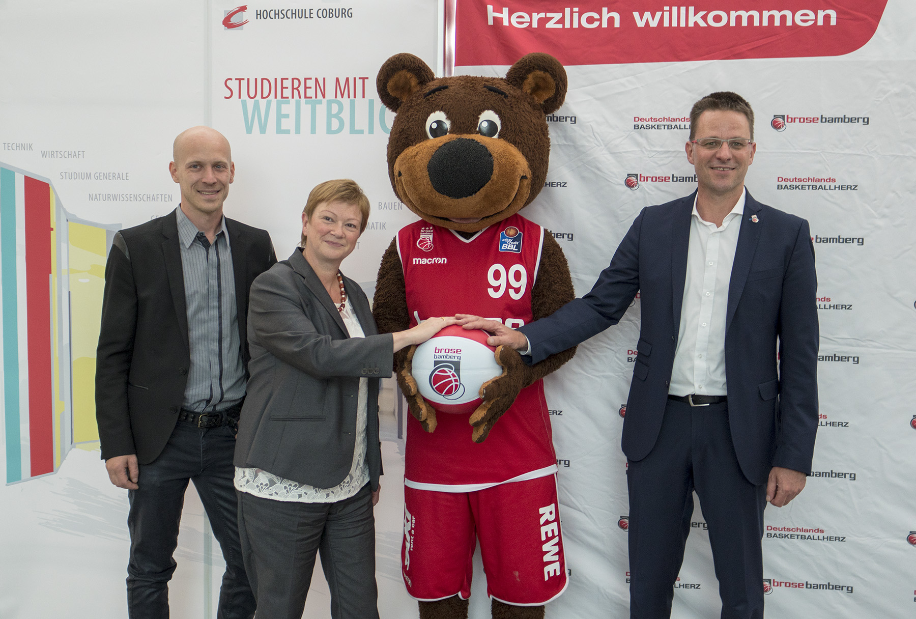 Drei Personen und ein Bären-Maskottchen posieren gemeinsam, jeder hält einen Basketball mit dem „Brose Bamberg“-Logo. Auf einem Banner dahinter steht „Herzlich willkommen“ der Hochschule Coburg und feiert das Thema „Studieren mit Weitblick“.