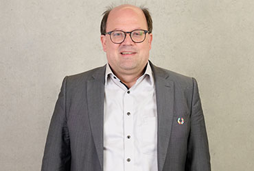 A person wearing glasses, a white shirt, and a gray suit jacket stands against a plain light-colored background. They have short hair and are smiling warmly. A small circular Hochschule Coburg pin adorns their jacket lapel.