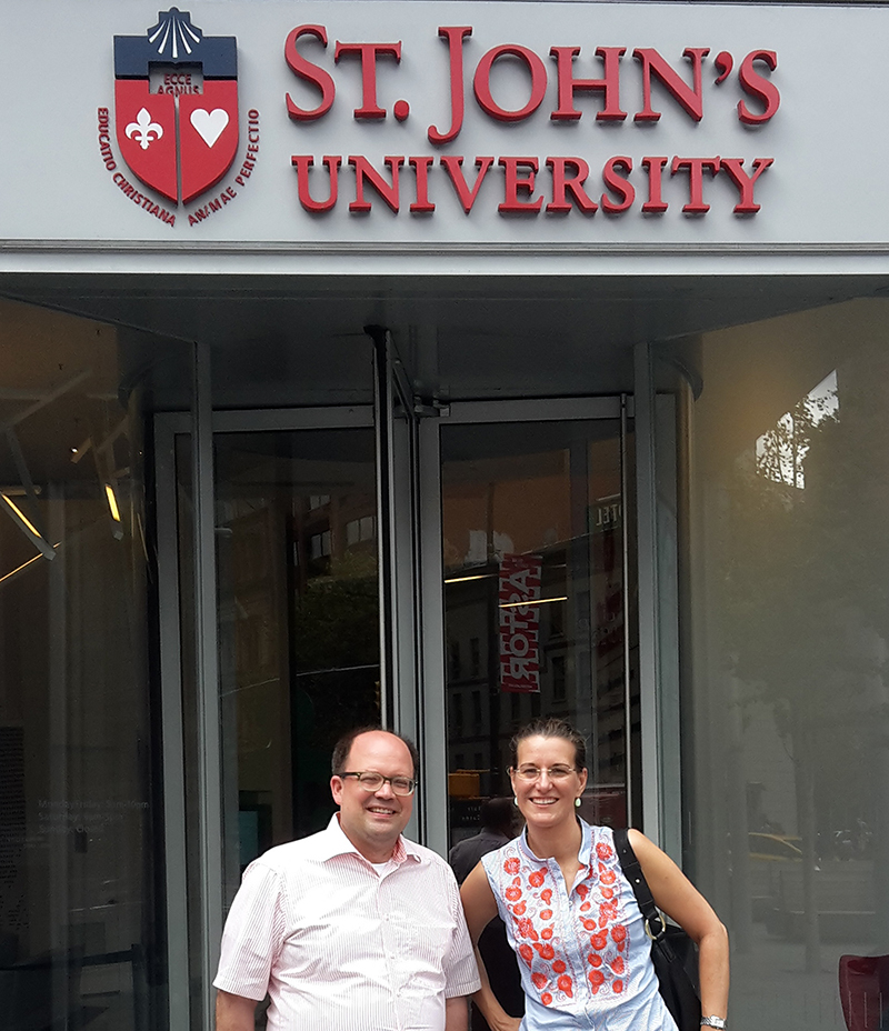 Zwei Menschen stehen lächelnd vor dem Eingang der St. John's University und erinnern damit an die einladende Atmosphäre der Hochschule Coburg. Über den Glastüren ist das Logo der Hochschule zu sehen. Die Person links trägt ein helles Hemd, die Person rechts eine gemusterte Bluse.