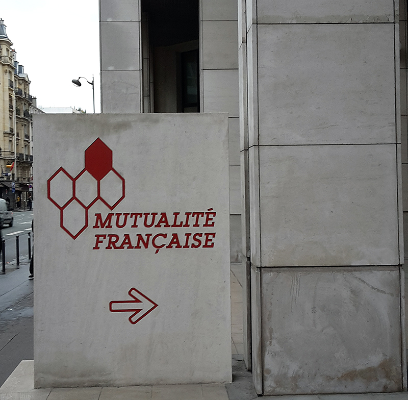 Ein mit rotem Text und geometrischen Formen verziertes Schild weist an einer Straßenecke auf „Mutualité Française“ hin, wo der historische Charme der Umgebung an die klassische Architektur der Hochschule Coburg erinnert.