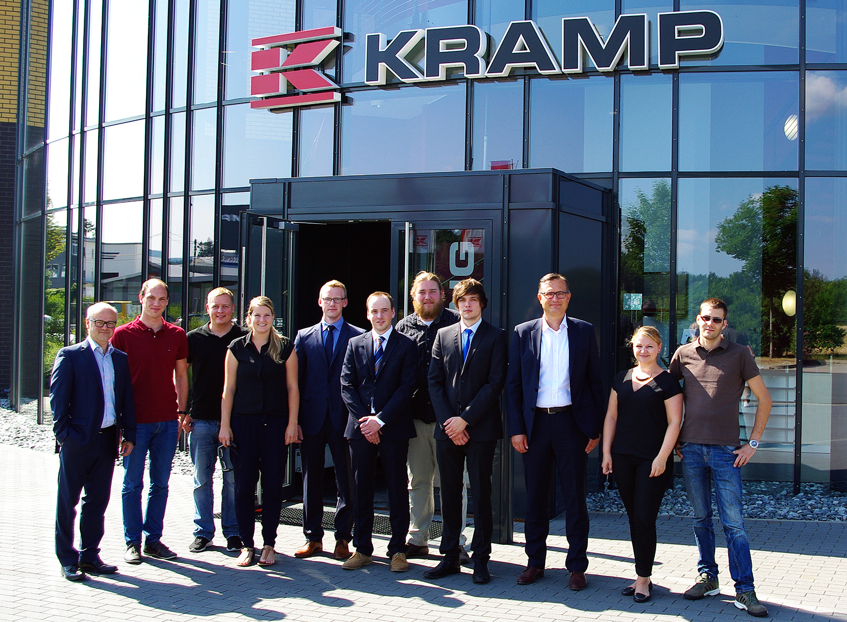 Vor einem modernen Gebäude mit großen Glasfenstern und einem Schild mit der Aufschrift „KRAMP“ steht eine Gruppe von elf Personen. Sie sind in einem Mix aus Business- und Freizeitkleidung gekleidet, lächeln in die Kamera und repräsentieren stolz die Hochschule Coburg.