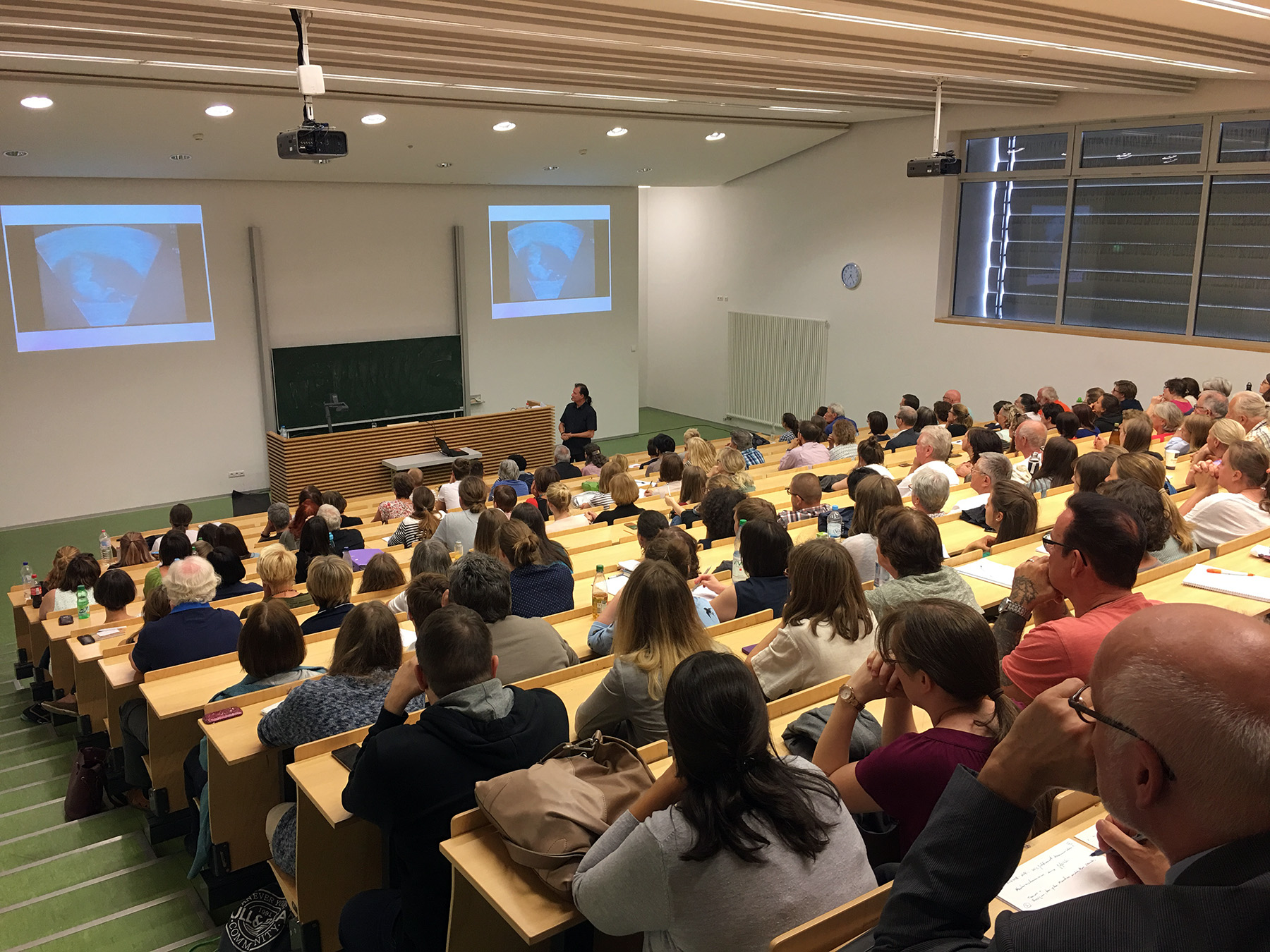 Im voll besetzten Hörsaal der Hochschule Coburg hält ein Redner einen Vortrag vor einem aufmerksamen Publikum. Dank der abgestuften Holzsitze und zwei Bildschirmen, auf denen das gleiche Bild gezeigt wird, können sich die Teilnehmer in einem hellen, modernen Ambiente auf die Präsentation konzentrieren.