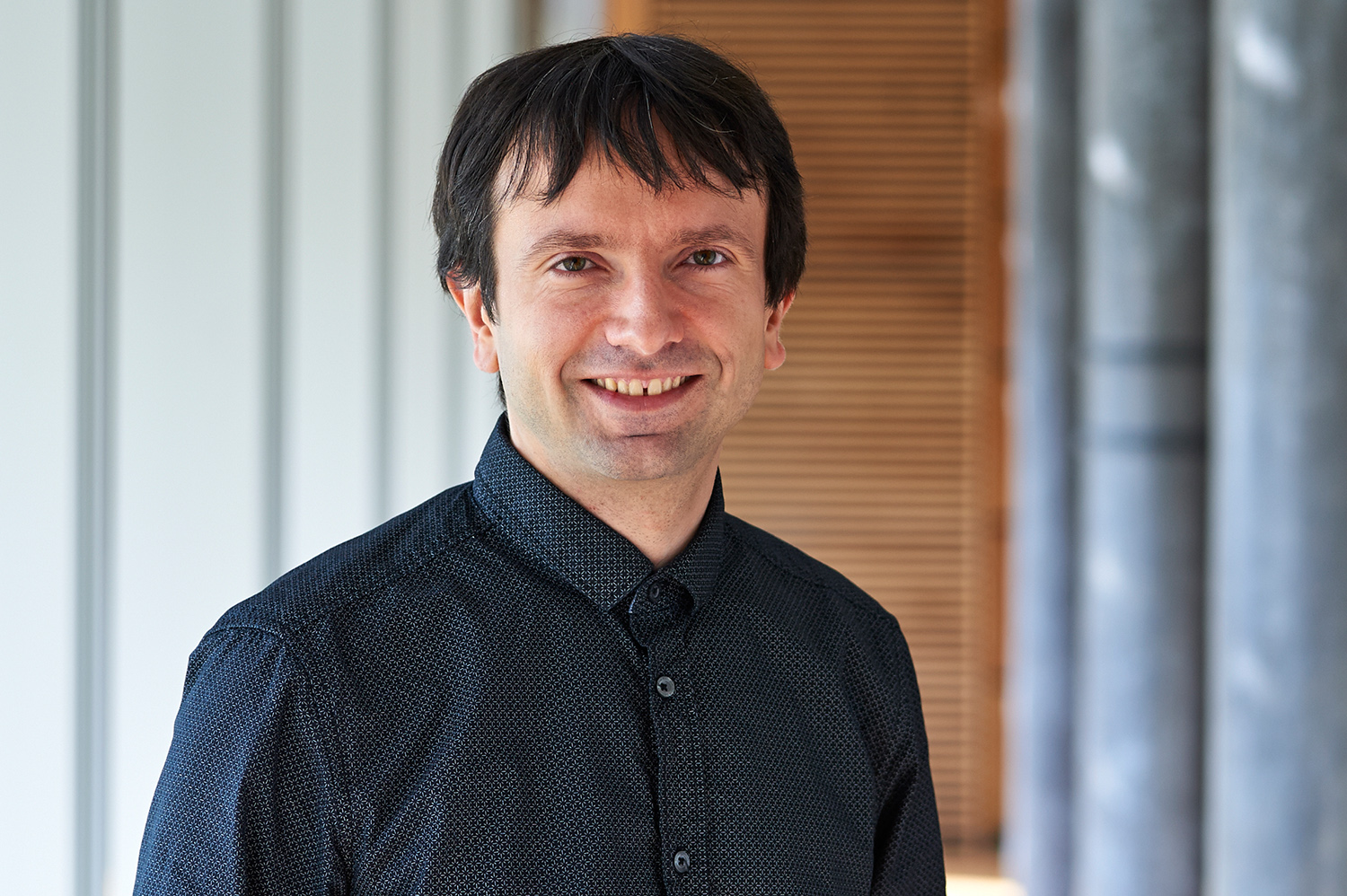 Eine Person mit kurzen dunklen Haaren lächelt und trägt ein dunkles Hemd mit Knöpfen. Der Hintergrund, der an die Architektur der Hochschule Coburg erinnert, besteht aus vertikalen Linien und Säulen, die der Szene eine akademische Eleganz verleihen.