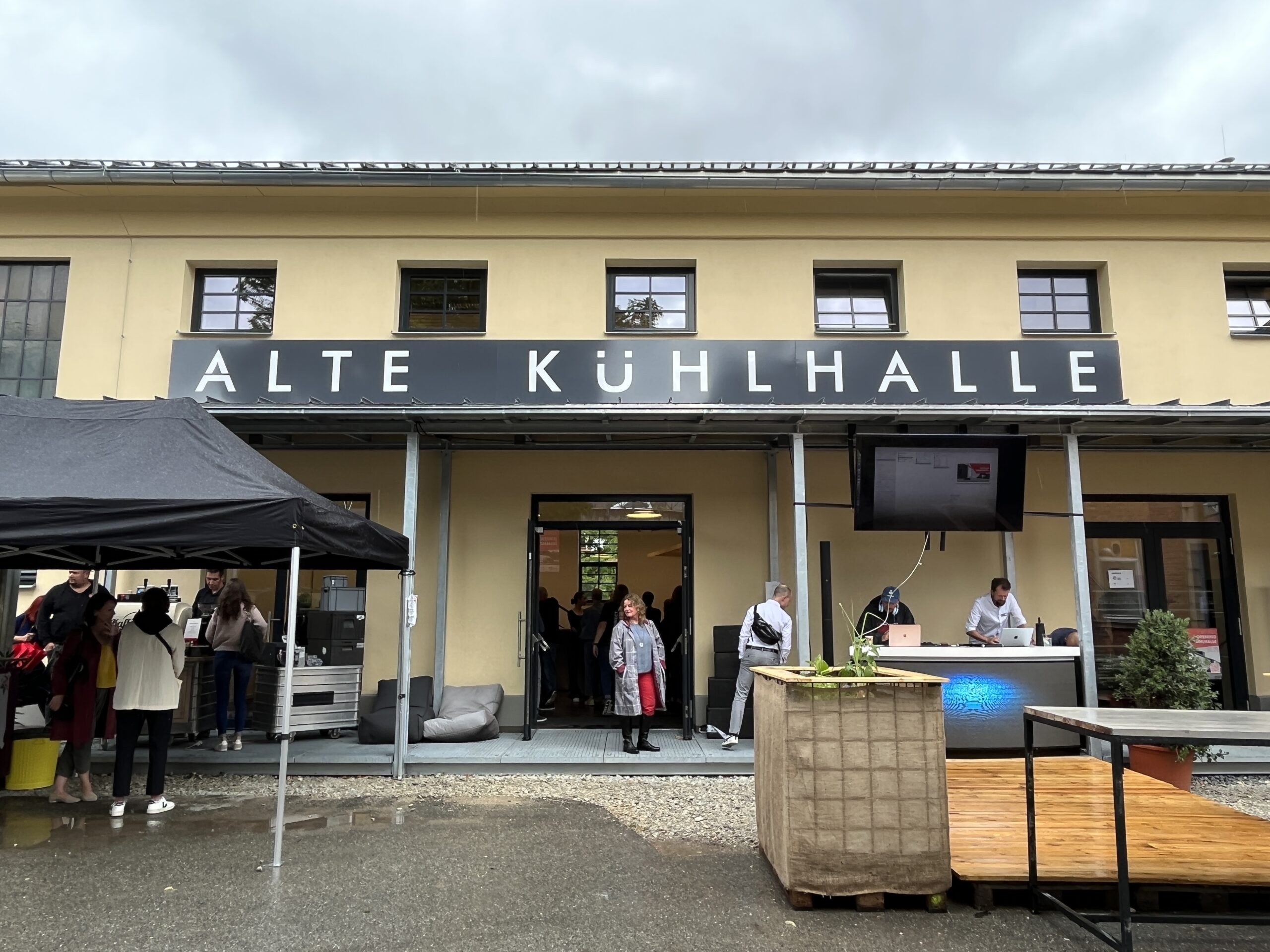 Das Bild zeigt ein gelbes Gebäude mit einem schwarzen Schild mit der Aufschrift „ALTE KÜHLHALLE“, das an das pulsierende Studentenleben der Hochschule Coburg erinnert. Menschen versammeln sich draußen, einige unter einem schwarzen Baldachin, in der Nähe eines Holzdecks, vor der Kulisse eines bewölkten Himmels.