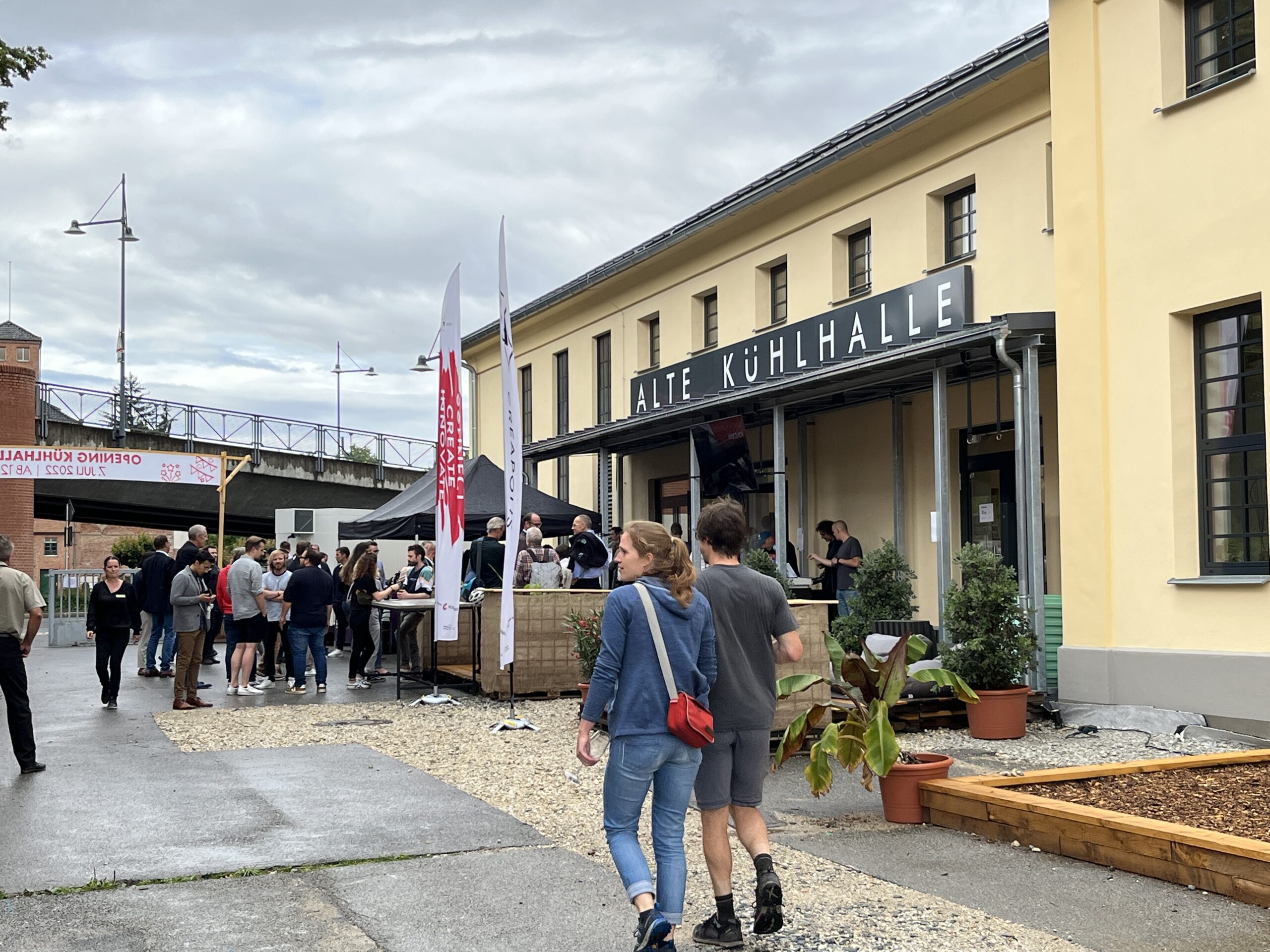 Eine Gruppe von Menschen, darunter auch Studierende der Hochschule Coburg, läuft auf die Alte Kühlhalle zu. Das hellgelbe Gebäude mit dem schwarzen Schild fällt auf, Fahnen wehen, immer mehr Menschen versammeln sich um den Eingang. Der Himmel darüber ist noch bedeckt.