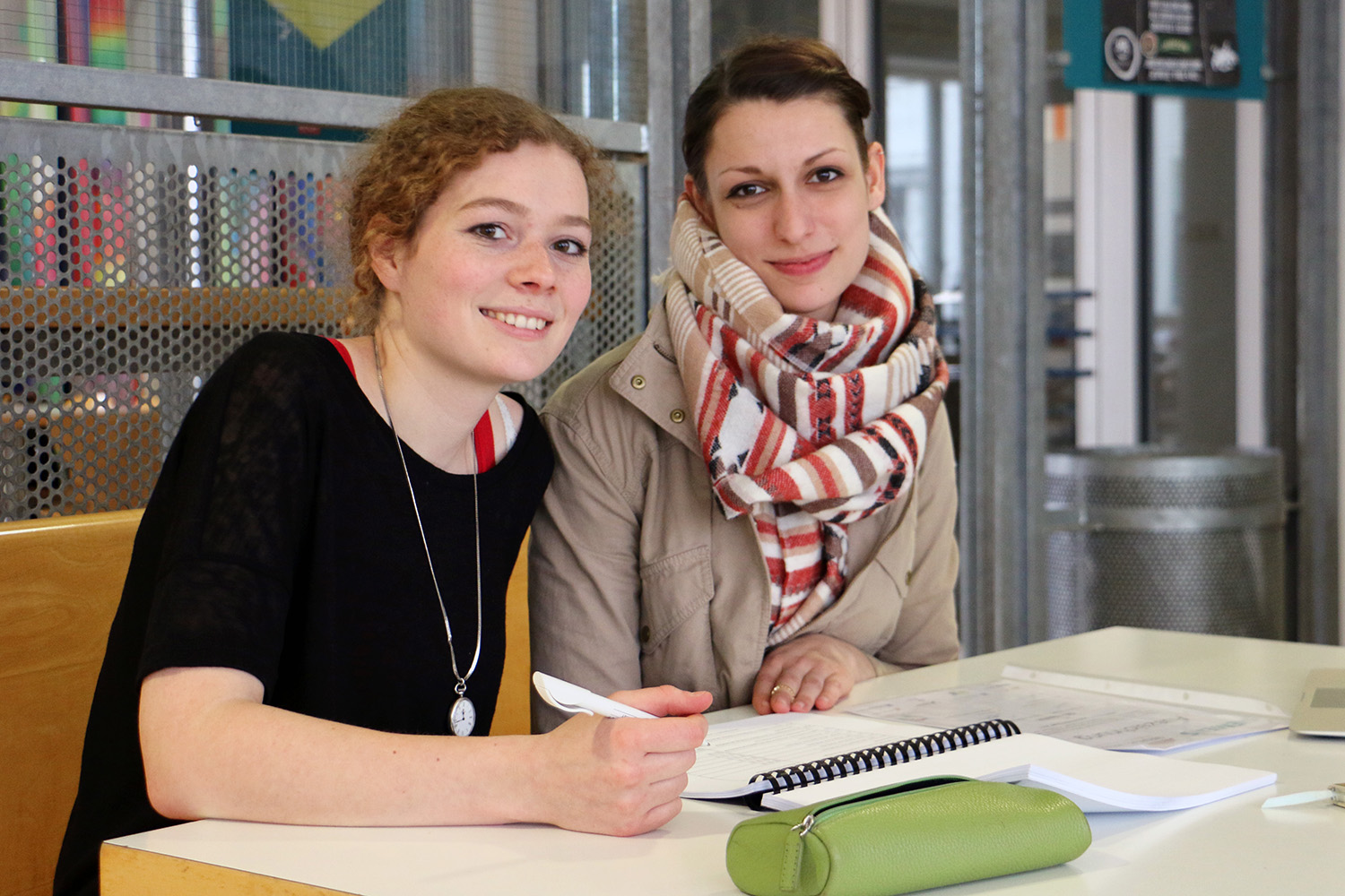 Zwei Frauen lächeln an einem Tisch in einem hellen Innenraum. Eine mit lockigem Haar hält einen Stift über ihrem Notizbuch, während die andere mit zurückgebundenem braunem Haar und gestreiftem Schal ihr gegenüber sitzt. Auf dem Tisch liegt ein grünes Federmäppchen, das die lebendige Atmosphäre der Hochschule Coburg einfängt.