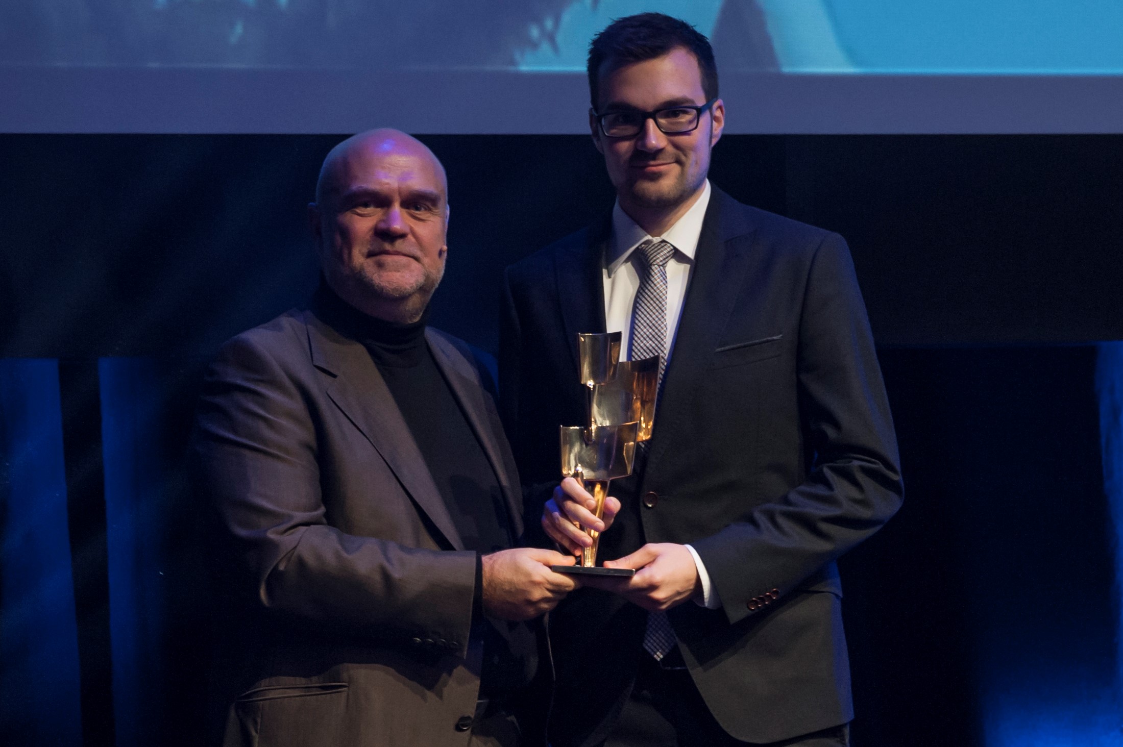 Bei der Veranstaltung der Hochschule Coburg stehen zwei Männer in Anzügen auf einer Bühne und halten gemeinsam eine goldene Trophäe in den Händen. Der Hintergrund ist blau und dunkel, was auf eine formelle Preisverleihung schließen lässt. Der Mann links ist kahl und der Mann rechts trägt eine Brille.