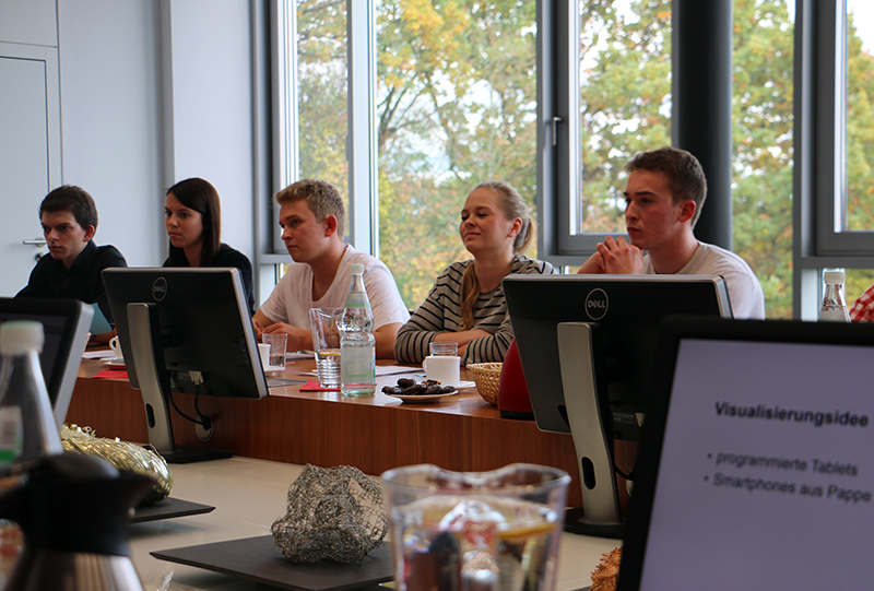 Eine Gruppe von Menschen sitzt an einem Konferenztisch in der Hochschule Coburg, tippt auf Computern und macht sich Notizen. Durch die großen Fenster des Raums blickt man auf die Bäume draußen, während im Vordergrund ein Bildschirm deutschen Text anzeigt.