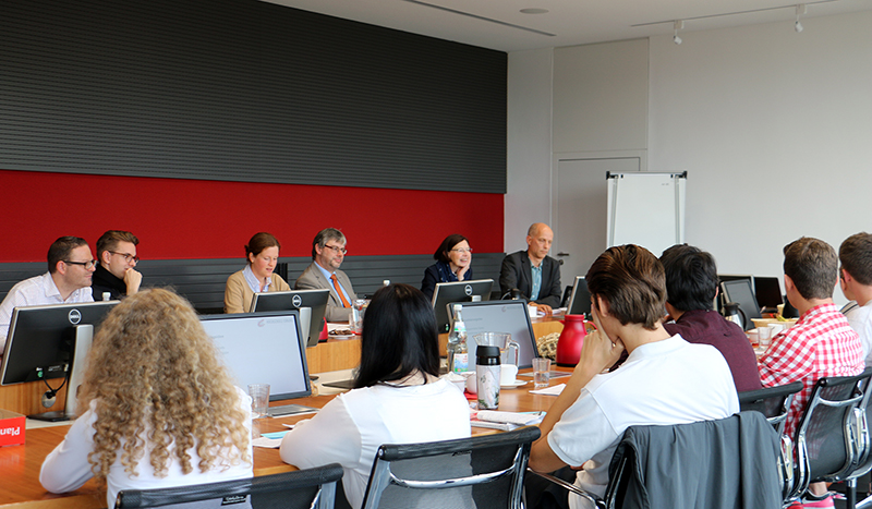 Eine Gruppe von Menschen sitzt um einen großen Konferenztisch in der Hochschule Coburg und nimmt an einer Besprechung teil. Mehrere Personen sitzen vor einem Rednerpult vor ihren Laptops. In der Nähe im Raum steht ein Whiteboard.