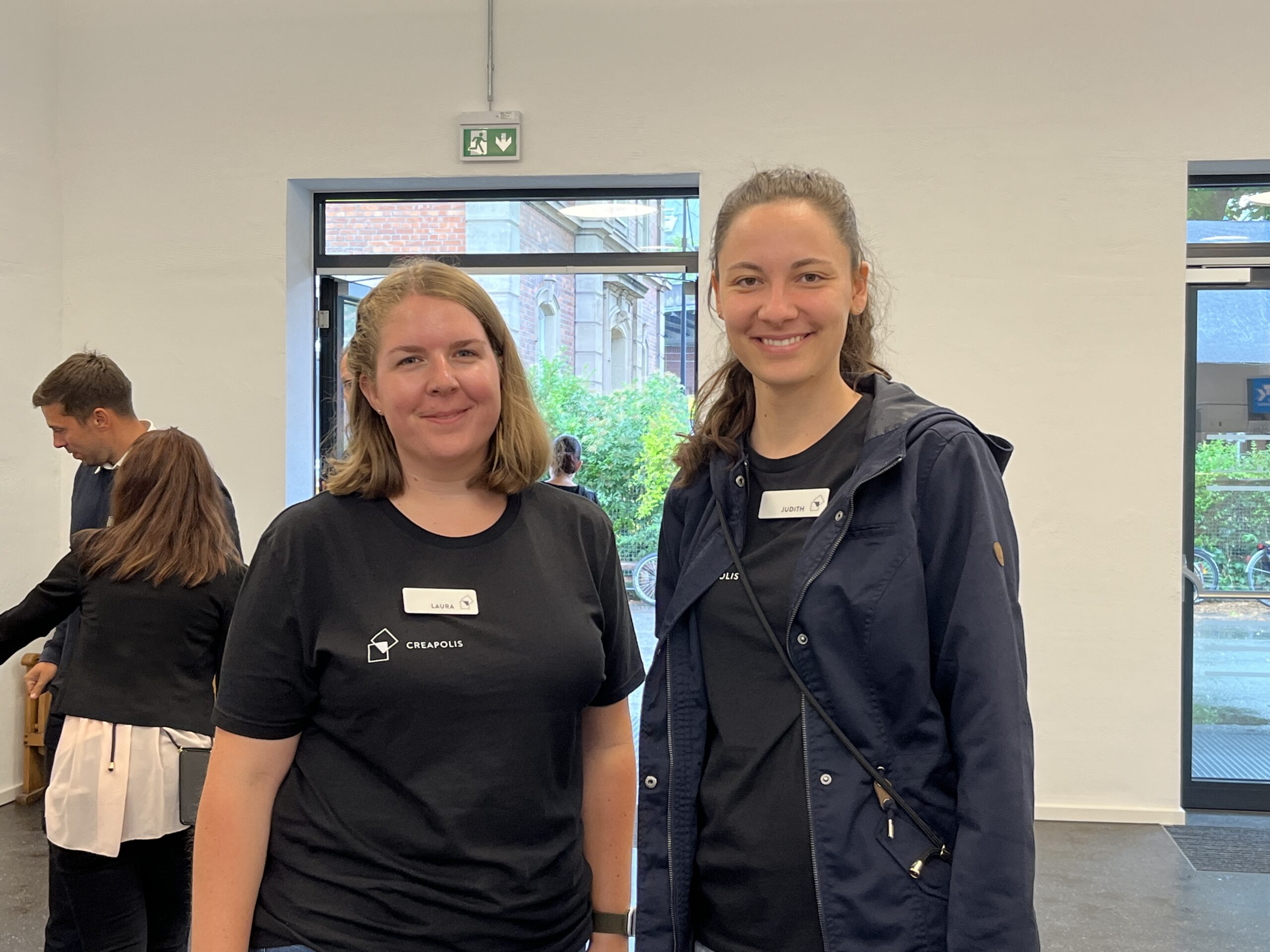 Zwei Frauen stehen drinnen und lächeln in die Kamera. Sie tragen schwarze Shirts mit Namensschildern und scheinen sich auf einer Veranstaltung der Hochschule Coburg zu befinden. Im Hintergrund sind weitere Personen in der Nähe offener Türen zu sehen, was zur lebhaften Atmosphäre beiträgt.