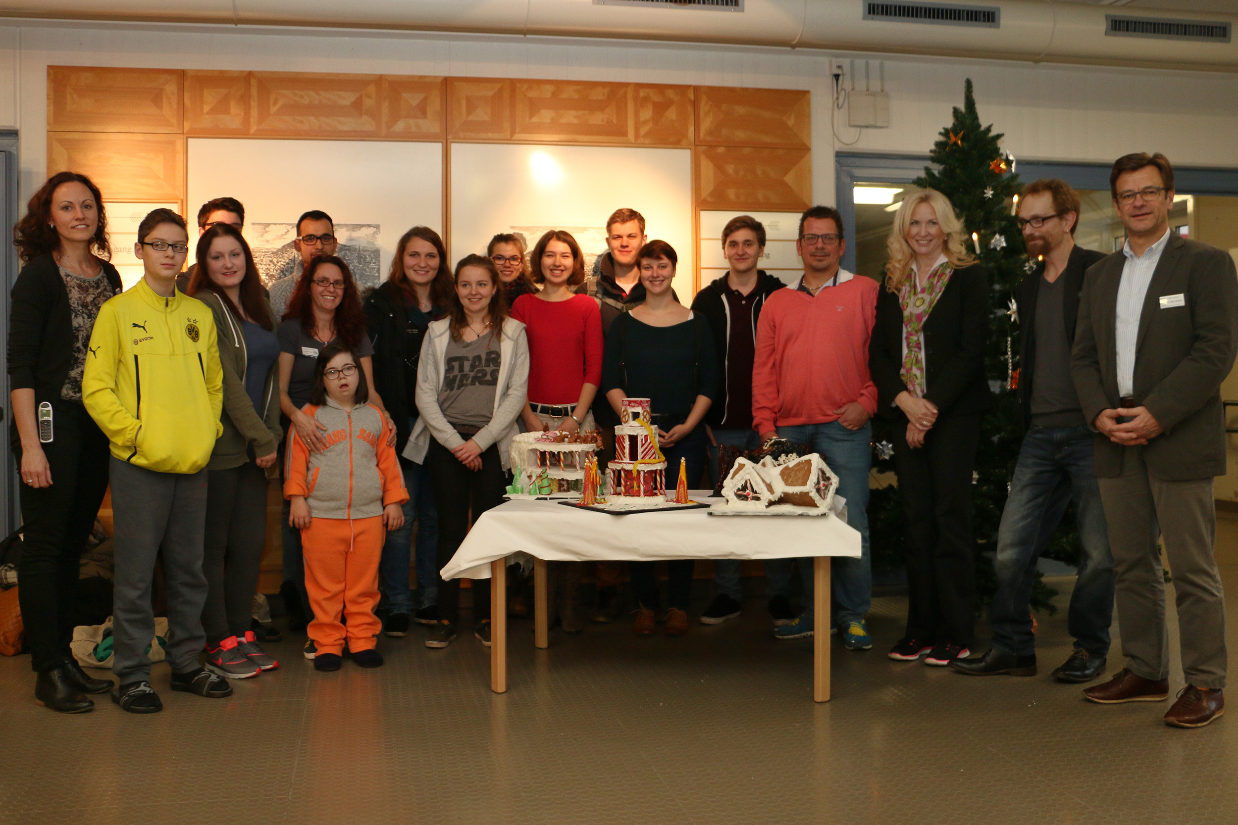 Eine Gruppe von Menschen, darunter Kinder und Erwachsene, versammelt sich um einen Tisch mit Lebkuchenhäusern. In dem festlichen Raum mit Weihnachtsbaum und teilweise sichtbarer Holzwand erinnert die Stimmung an eine fröhliche Werkstatt an der Hochschule Coburg.