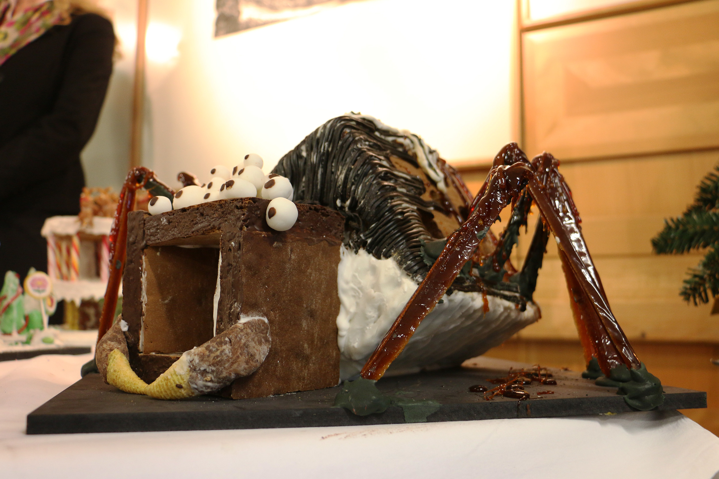 Ein Lebkuchenhaus in Form einer riesigen Spinne mit Schokoladengliedern und einem Körper aus weißem Zuckerguss. Mit Zuckeraugen und anderen Süßigkeiten dekoriert, wird es als Teil der festlichen Ausstellung im Innenbereich der Hochschule Coburg auf einem Tisch ausgestellt.