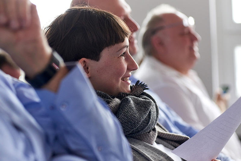 Eine Person mit kurzen Haaren und Kopftuch sitzt lächelnd in einer Reihe von Menschen bei einer Indoor-Veranstaltung der Hochschule Coburg. Sie hält ein Blatt Papier in der Hand und auch andere neben ihr sind engagiert. Der Fokus liegt auf dem fröhlichen Ausdruck.