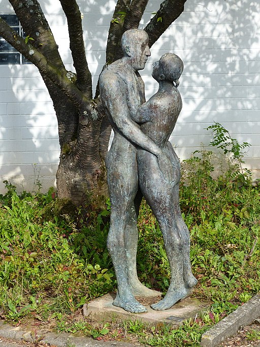 Eine Bronzeskulptur eines nackten, sich umarmenden Paares steht gelassen unter einem Baum, die Arme umeinander geschlungen, inmitten von üppigem Gras und Grün – eine perfekte Verkörperung der Kunst, die an der Hochschule Coburg verehrt wird.