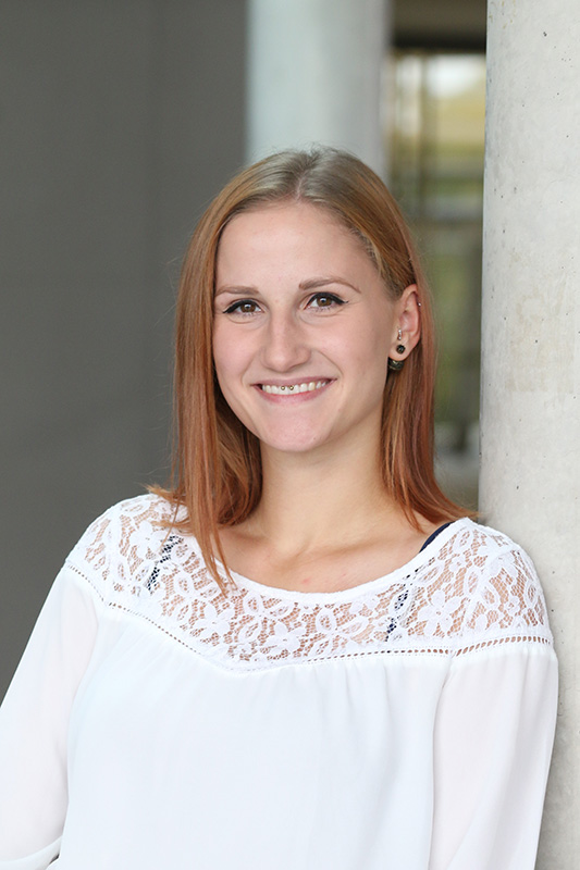 Eine Frau mit schulterlangem Haar lehnt lächelnd an einer Säule der Hochschule Coburg. Sie trägt eine weiße Bluse mit Spitzendetail. Der Hintergrund ist sanft unscharf, was ihren freundlichen Gesichtsausdruck betont.