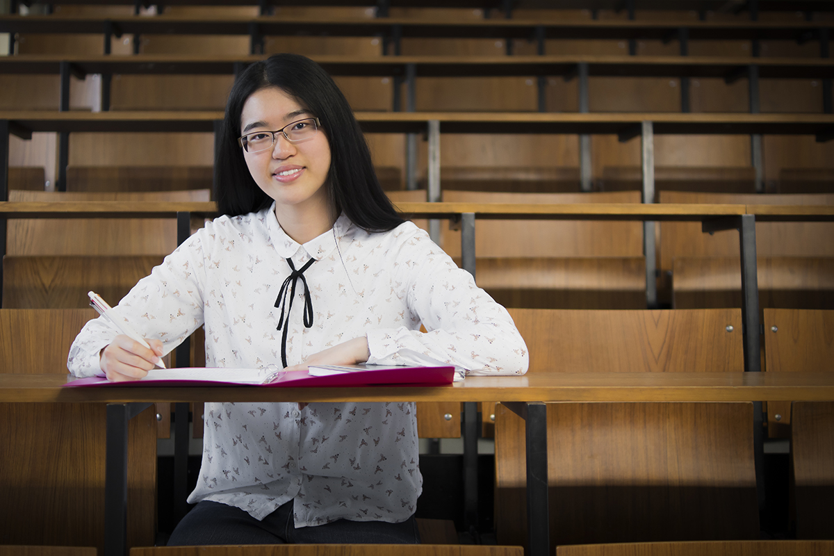 Inmitten der leeren Holzbänke im Hörsaal der Hochschule Coburg sitzt eine Frau mit langen schwarzen Haaren und Brille allein und schreibt in ein rosa Notizbuch. Ihre weiße Bluse, die mit einem dezenten Muster verziert ist, bildet einen sanften Kontrast zur akademischen Einsamkeit, die sie umgibt.