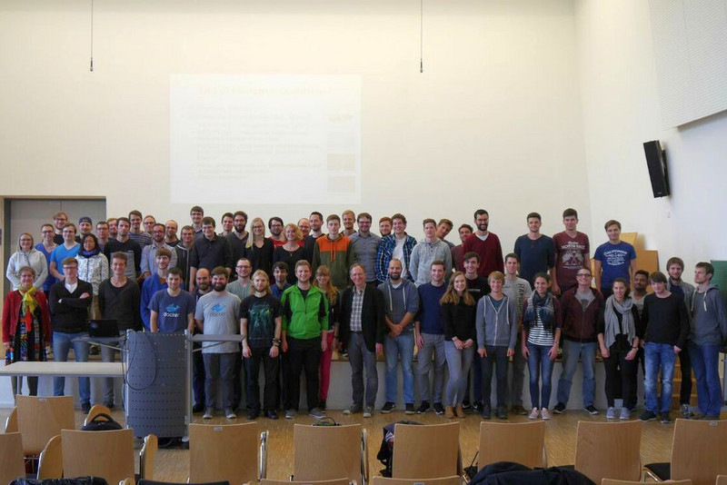 Eine große Gruppe von Menschen, darunter Männer und Frauen, steht in einem Hörsaal der Hochschule Coburg. Sie posieren für ein Foto vor einer Präsentationsleinwand. Einige lächeln, und man sieht unterschiedliche Freizeitkleidung. Im Vordergrund sind leere Stühle zu sehen.