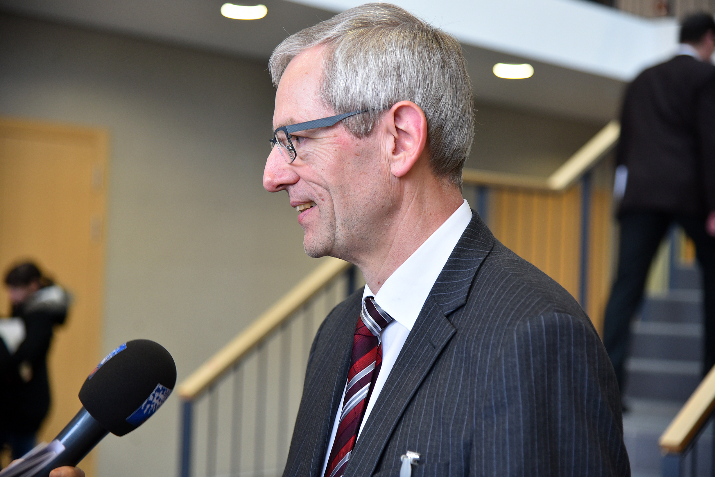 Ein Mann in Anzug und Brille wird in einem Innenbereich der Hochschule Coburg interviewt. Er lächelt und hält ein schwarzes Mikrofon in der Hand. Im Hintergrund ist eine Treppe zu sehen.