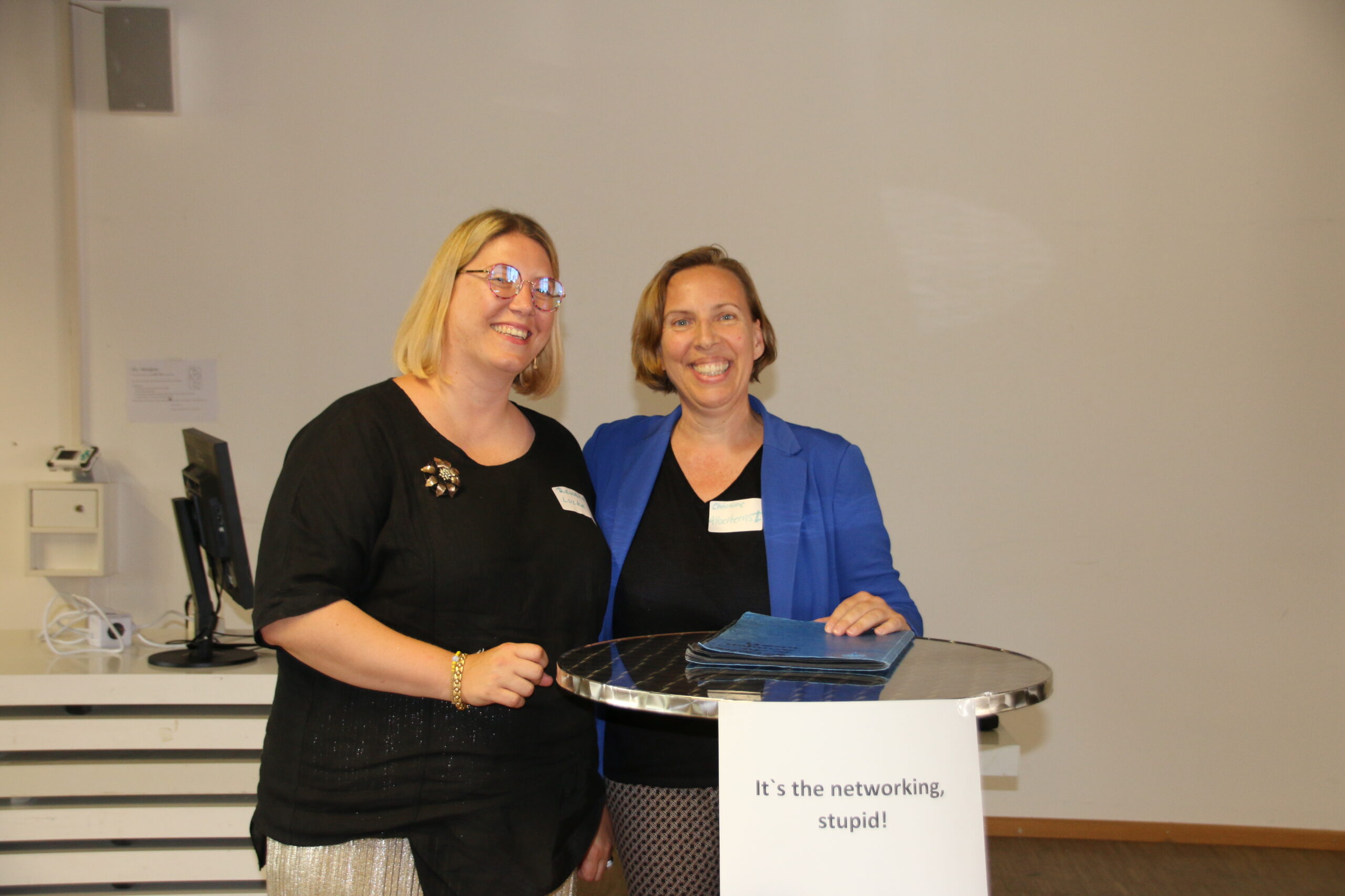 Zwei lächelnde Frauen stehen hinter einem Podium mit einem Schild mit der Aufschrift „It’s the networking, stupid!“ an der Hochschule Coburg. Sie sind drinnen, eine Frau trägt ein schwarzes Hemd und die andere einen blauen Blazer. Auf dem Podium liegt ein Stapel Ordner oder Papiere.