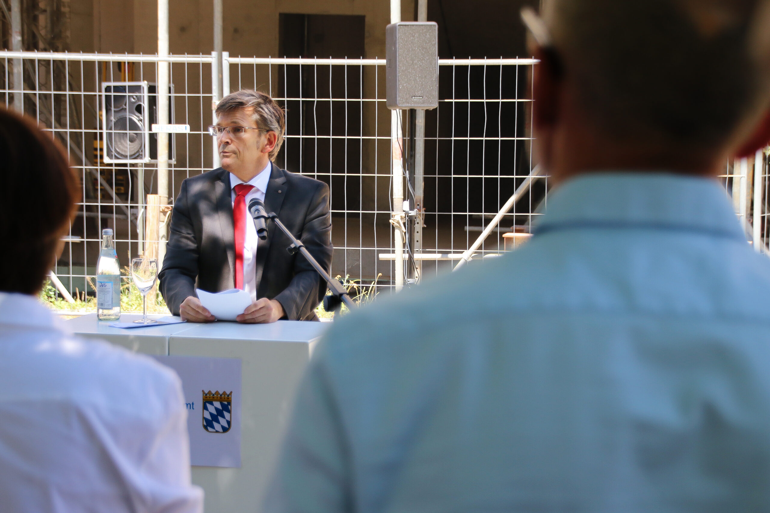 Ein Mann im Anzug spricht an einem Podium im Freien und hält einen Zettel mit dem Emblem der Hochschule Coburg in der Hand. Auf dem Podium steht ein Mikrofon neben dem bayerischen Wappen. Aufmerksame Zuhörer füllen den Vordergrund, im Hintergrund ein Metallzaun und ein Rednerpult.