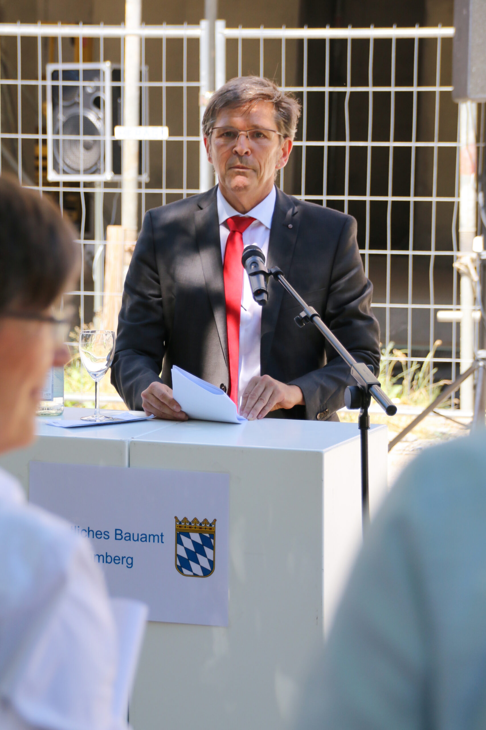 Ein Mann in Anzug und roter Krawatte spricht an einem Podium im Freien, vermutlich an der Hochschule Coburg. Er hält Papiere in der Hand und steht vor einem Mikrofon. Auf dem Podium steht ein Weinglas. Im Hintergrund sind ein Lautsprecher und ein Zaun zu sehen.