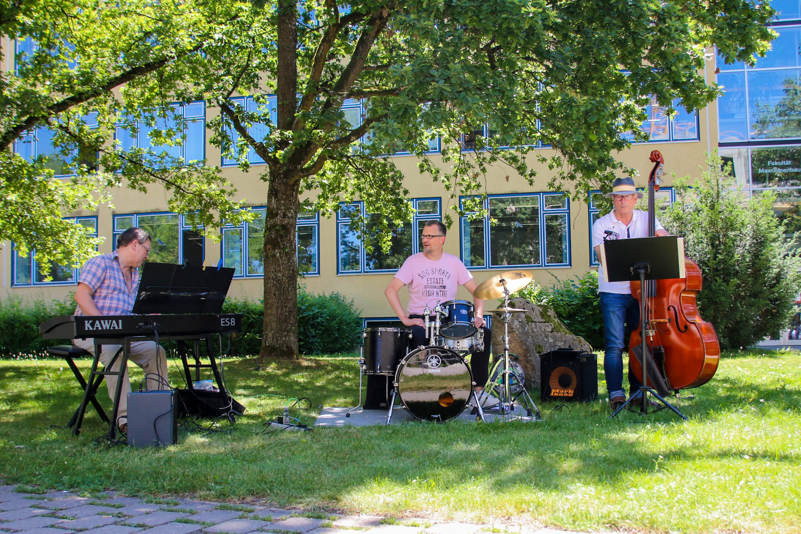 Drei Musiker spielen im Freien auf einer Rasenfläche der Hochschule Coburg. Einer spielt Keyboard, ein anderer Schlagzeug und der dritte Kontrabass. Im Hintergrund lugen Bäume und ein Gebäude mit Fenstern hervor, alles unter einem sonnigen Himmel.