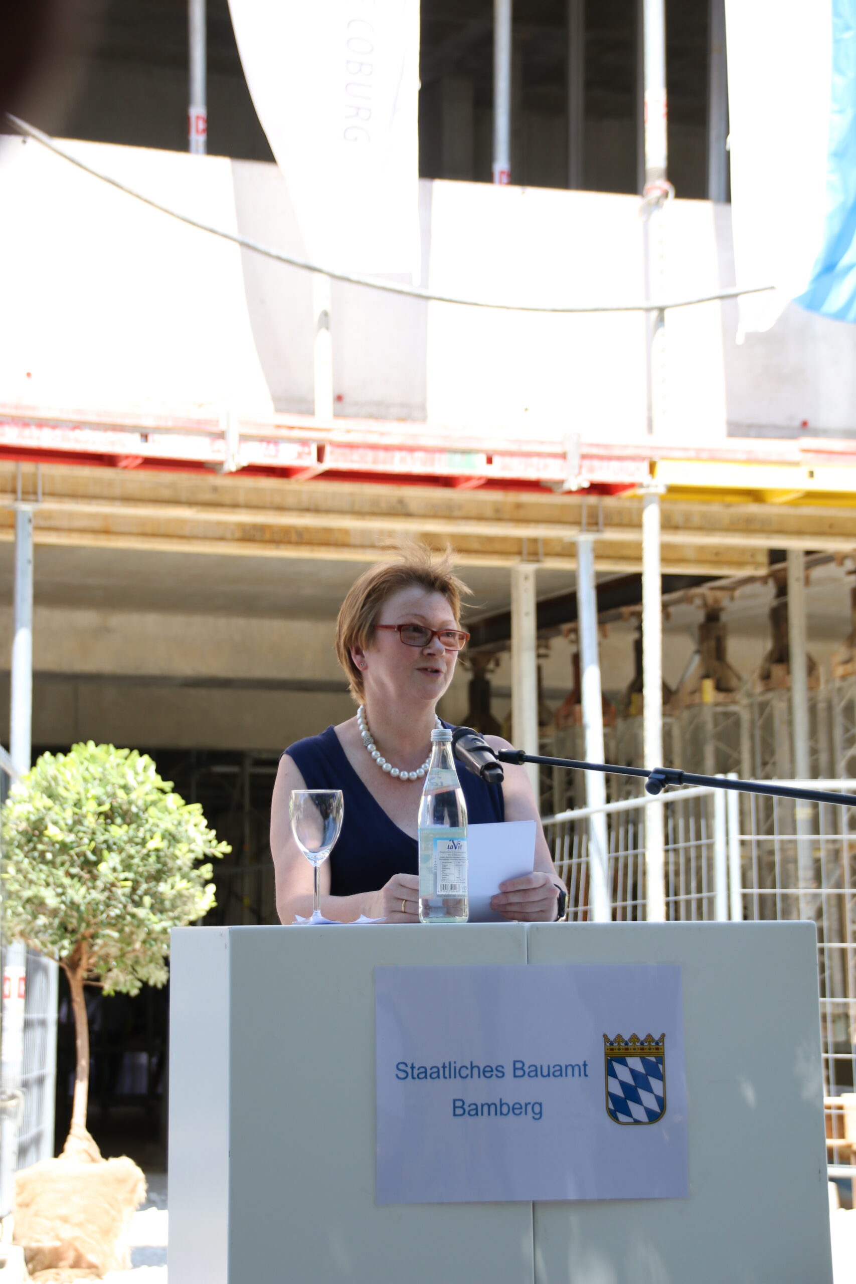 Eine Person steht an einem Podium, sie repräsentiert die Hochschule Coburg und spricht in ein Mikrofon. Sie hält ein Stück Papier in der Hand und steht vor einer Baustelle. Auf dem Podium stehen eine Flasche Wasser und ein Glas, auf dem ein Schild mit der Aufschrift „Staatliches Bauamt Bamberg“ steht.