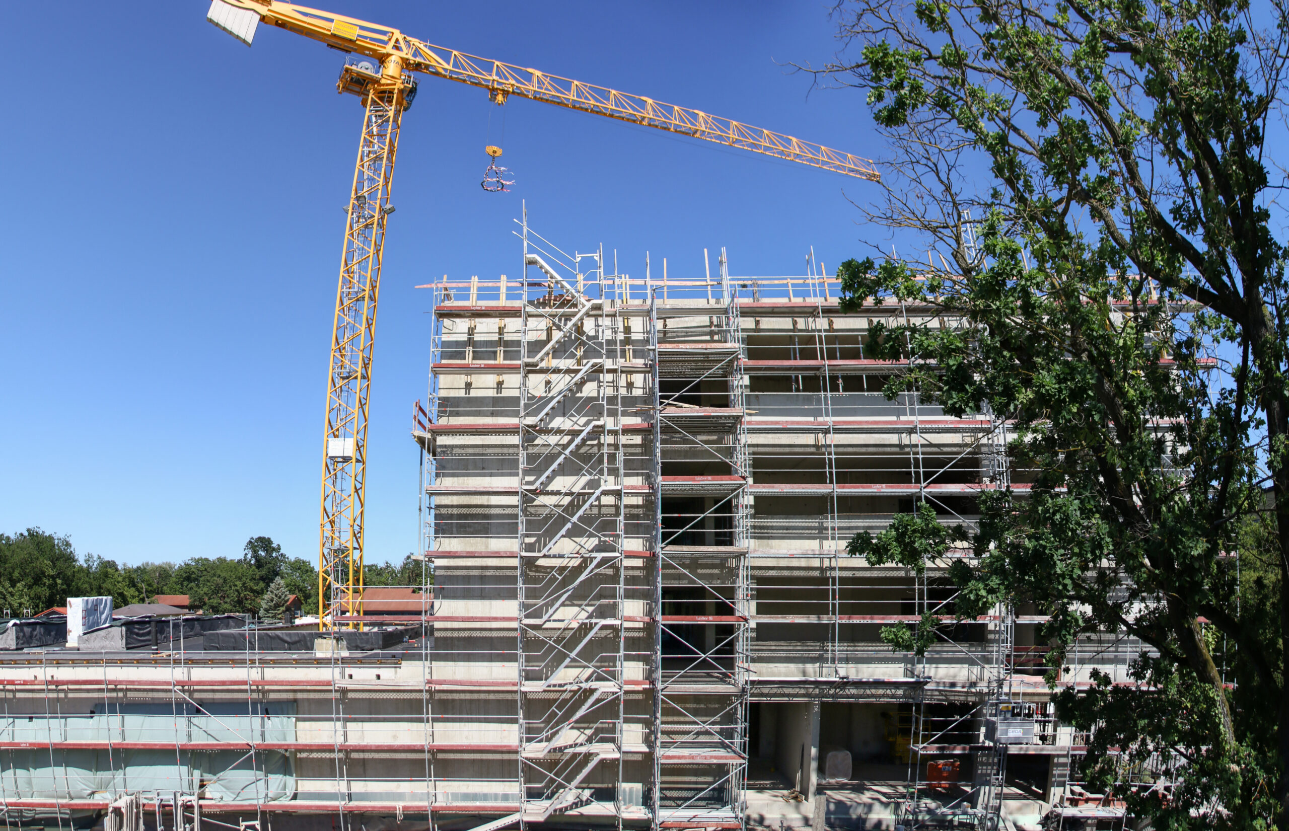 Auf einer Baustelle in der Nähe der Hochschule Coburg ist ein mehrstöckiges Gebäude mit Gerüst zu sehen. Ein hoher gelber Kran ragt über die Struktur hinaus, während rechts ein Baum vor dem klaren blauen Himmel steht.