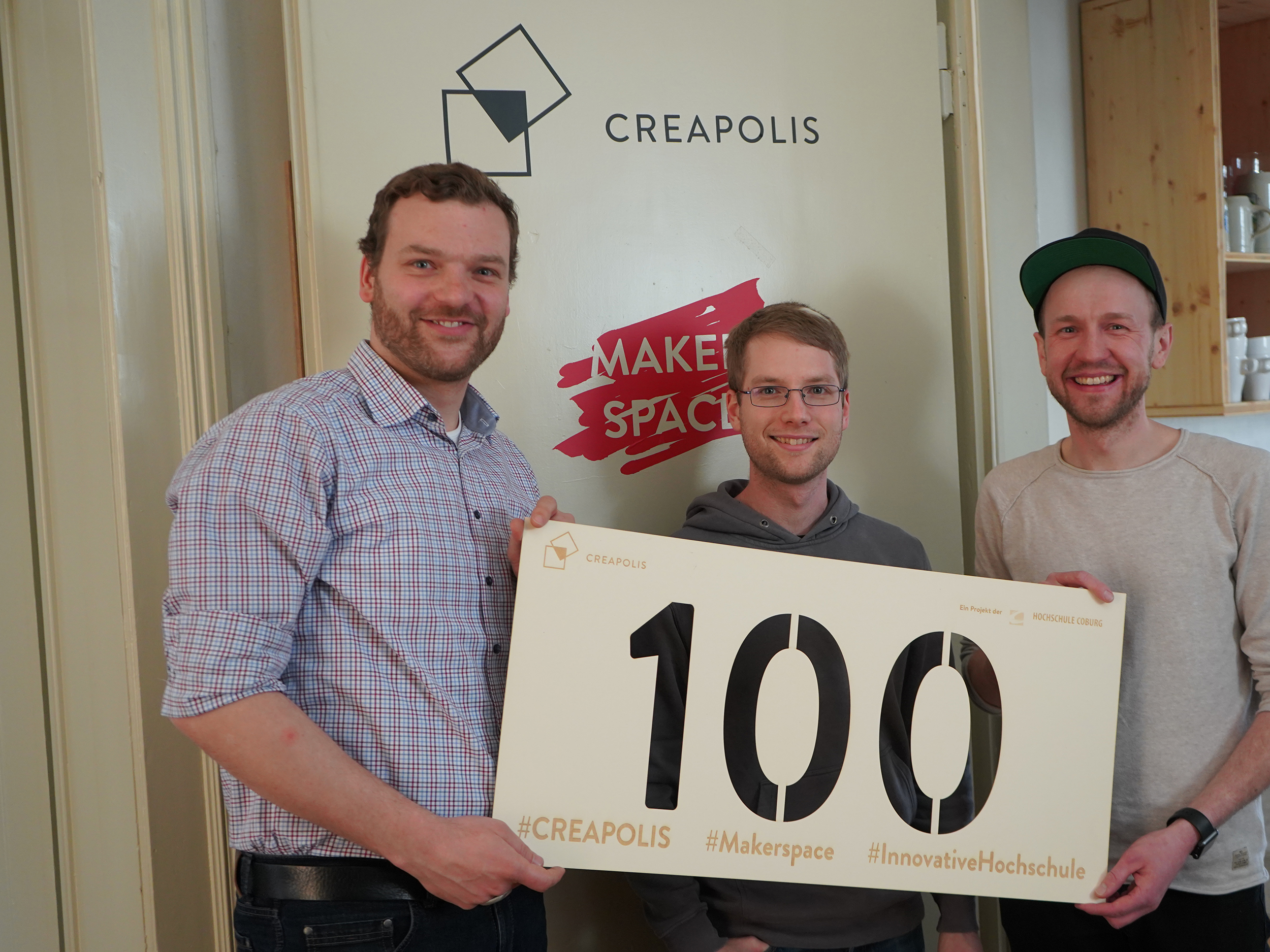 Drei Menschen stehen lächelnd und mit einem großen Schild mit der Zahl „100“ vor einer Tür mit der Aufschrift „CREAPOLIS“ und zeigen mit einem prominent angebrachten „Makerspace“-Aufkleber stolz ihre Verbundenheit zur Hochschule Coburg.