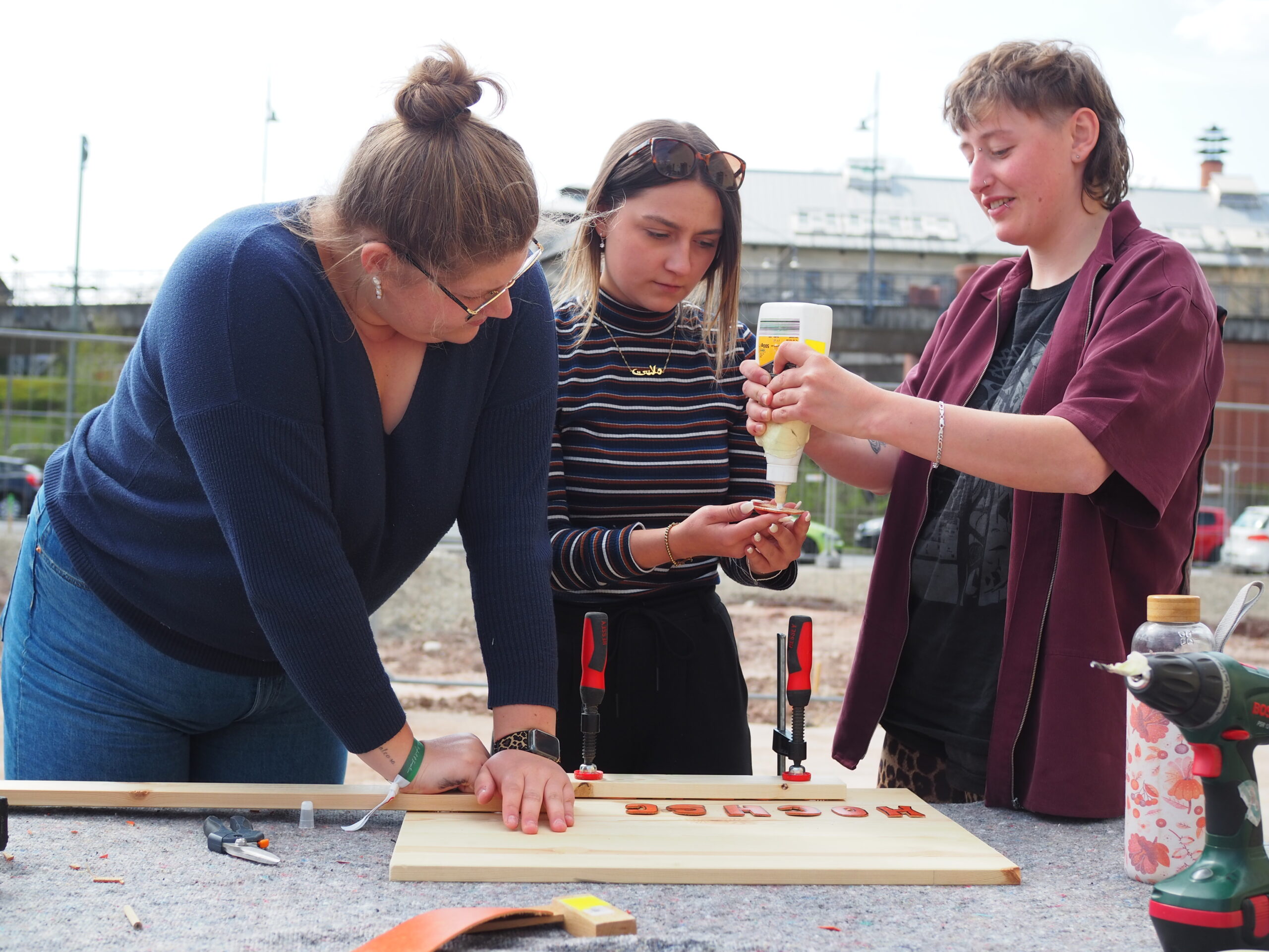 Drei Personen arbeiten gemeinsam an einem Holzbearbeitungsprojekt im Freien an der Hochschule Coburg. Sie verwenden Werkzeuge wie Klammern und Klebstoff und haben ein mit Buchstaben beschriftetes Stück Holz vor sich. Im Hintergrund sind Gebäude und ein Zaun unscharf zu sehen, was den Geist des praktischen Lernens einfängt.