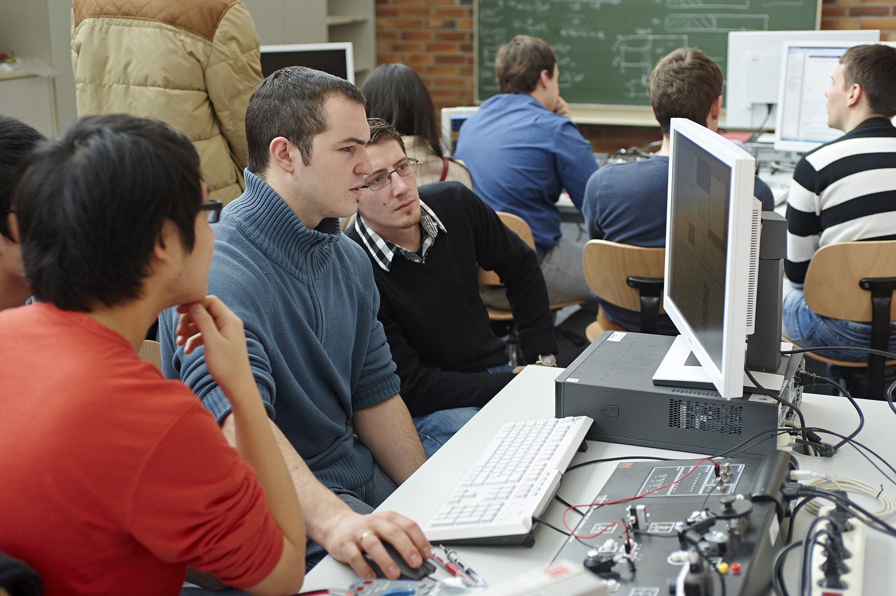 Eine Gruppe von Studierenden der Hochschule Coburg sitzt in einem Hörsaal an Schreibtischen mit Computern. Sie sind auf einen Bildschirm konzentriert und vertieft in ein gemeinsames Projekt. Im Hintergrund ragt eine mit Zeichnungen und Gleichungen geschmückte Tafel empor, die ihr akademisches Streben widerspiegelt.