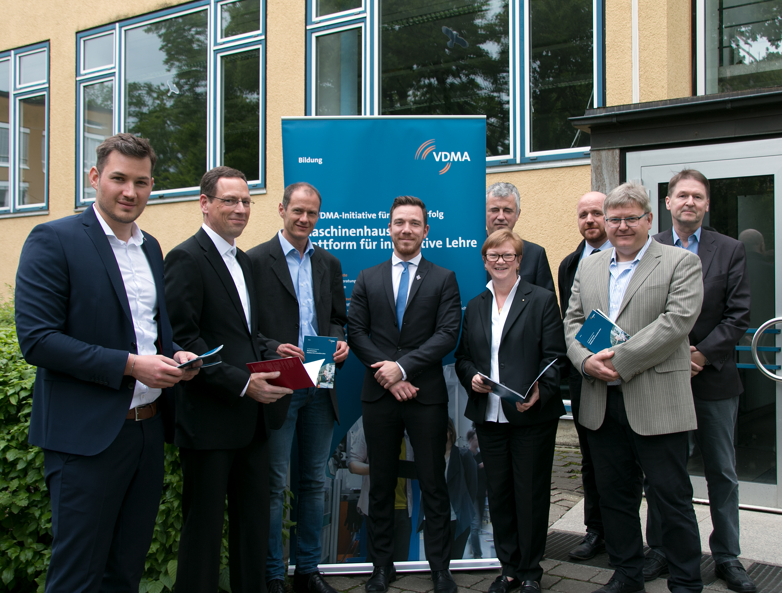 Eine Gruppe von Menschen in Business-Kleidung steht vor einem Gebäude mit einem VDMA-Banner, das die Hochschule Coburg repräsentiert. Sie halten Prospekte in den Händen und lächeln selbstbewusst in die Kamera.