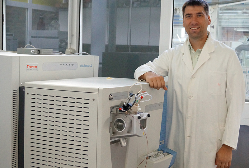 Ein Mann im Laborkittel steht lächelnd neben einem großen wissenschaftlichen Instrument an der Hochschule Coburg. Das weiße Gerät mit seinen zahlreichen Bedienfeldern und Displays fügt sich in die helle Laborumgebung ein, in der Glaswände und sichtbare Labormöbel für eine moderne Kulisse sorgen.