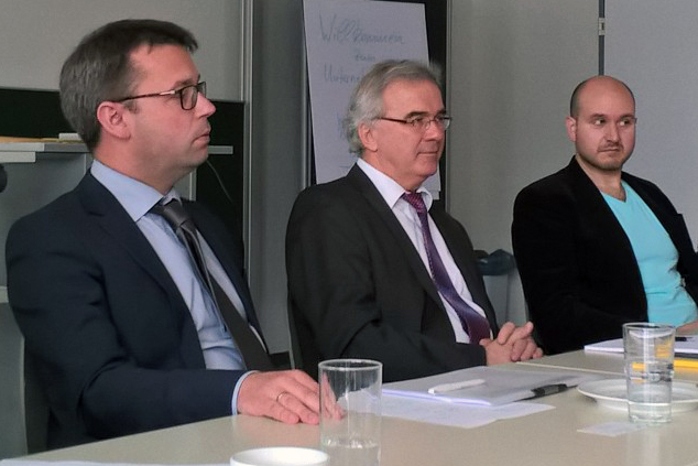 Drei Männer im Business-Outfit sitzen an einem Konferenztisch in der Hochschule Coburg. Zwei tragen Brillen, einer trägt ein hellblaues Hemd unter einem dunklen Blazer. Vor ihnen liegen Papiere und Gläser mit Wasser auf dem Tisch, im Hintergrund steht ein Flipchart.