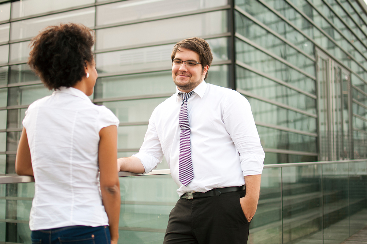 Zwei Menschen im Business-Outfit unterhalten sich im Freien nahe der modernen Glasfassade der Hochschule Coburg. Einer, in weißem Hemd und Krawatte, lehnt lässig an einem Geländer, die andere, in weißer Bluse, steht ihm gegenüber.