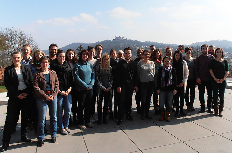 Eine Gruppe von Menschen steht auf einer Terrasse mit malerischer Aussicht auf sanfte Hügel und das Schloss der Hochschule Coburg in der Ferne. Sie sind leger gekleidet und lächeln, eingerahmt von Bäumen und einem klaren Himmel im Hintergrund.