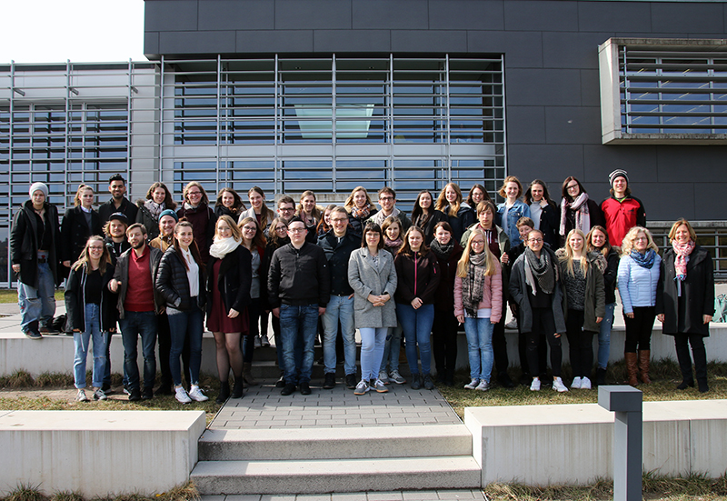 Eine große Gruppe von Menschen steht auf den Stufen vor einem modernen Gebäude der Hochschule Coburg. Sie sind leger und halbformell gekleidet, lächeln und posieren unter einem klaren Himmel für die Kamera.