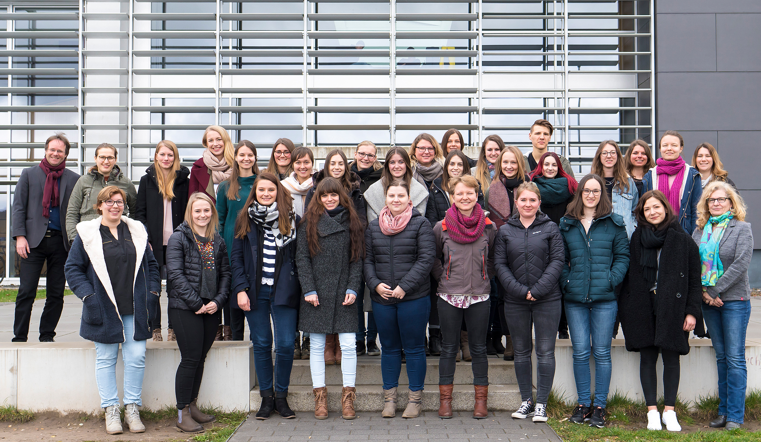 Eine Gruppe von Menschen posiert gemeinsam vor dem modernen Gebäude der Hochschule Coburg mit großen Fenstern. Sie tragen lässige Winterkleidung, darunter Mäntel und Schals. Das Wetter scheint kühl und bewölkt zu sein.