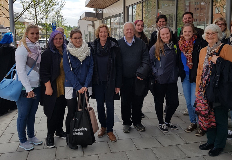 Eine Gruppe von Menschen steht zusammen auf einem Bürgersteig vor der Hochschule Coburg und posiert für ein Foto. Sie sind alle leger und warm gekleidet, und einige lächeln. Im Hintergrund sind Bäume und Gebäude zu sehen.